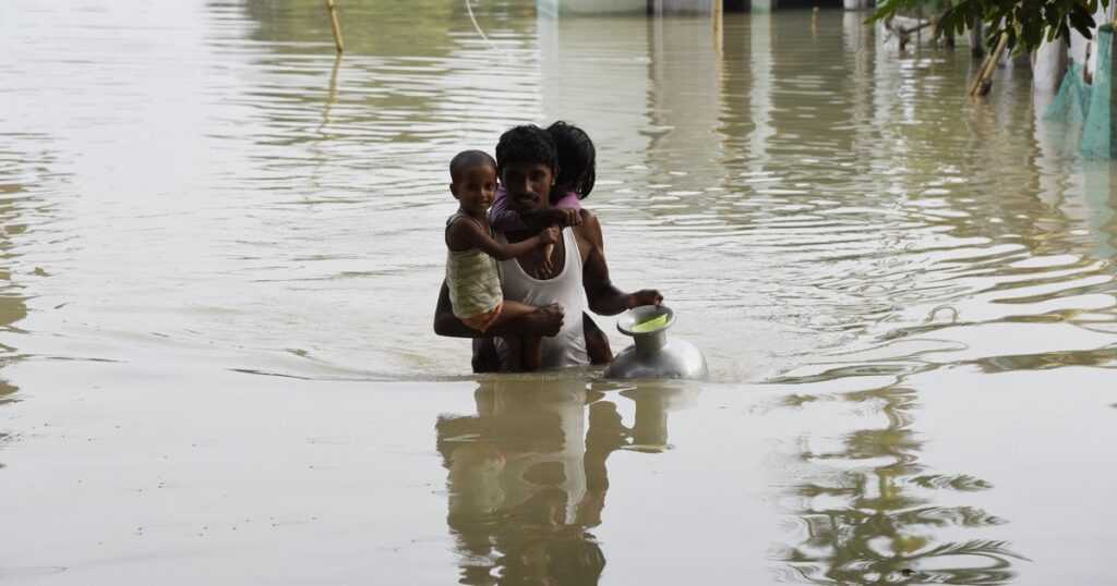 A father carries his two children in a waist-deep water that has swallowed his village in Assam (HuffPost)